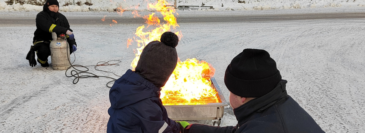 Lapsi suihkuttaa palosammuttimella tuleen palokuntalaisen avustuksella.